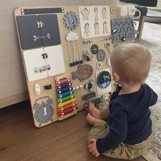 A parent and child sitting together on the floor, smiling and exploring a sensory board with locks and latches